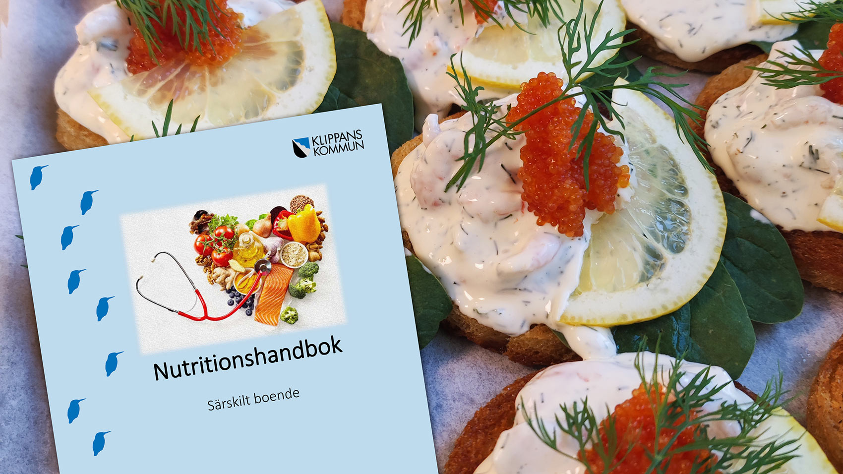 En närbild på festliga smörgåsar toppade med räkor, rom, dill och citronskivor, bredvid en bok från Klippans kommun med titeln "Nutritionshandbok – Särskilt boende".