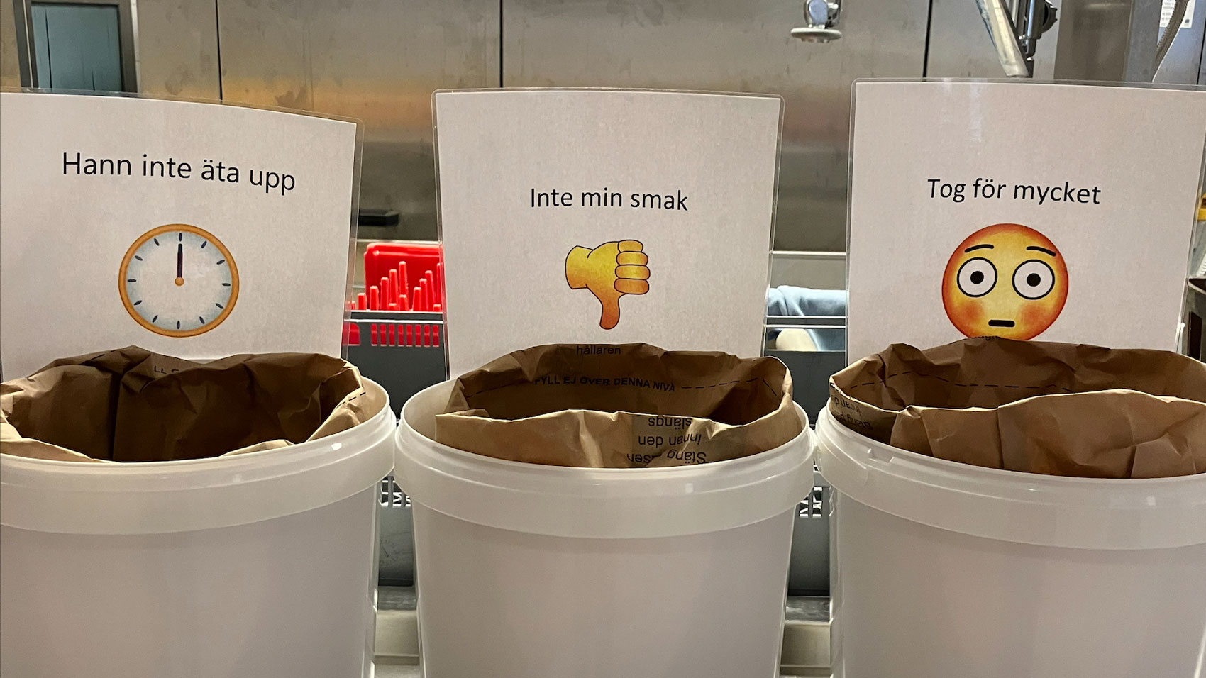Tre vita soptunnor märkta med skyltar för att indikera orsaker till matsvinn. Den första tunnan är märkt "Hann inte äta upp" med en klocka, den andra "Inte min smak" med en tummen ner, och den tredje "Tog för mycket" med en förvånad emoji.