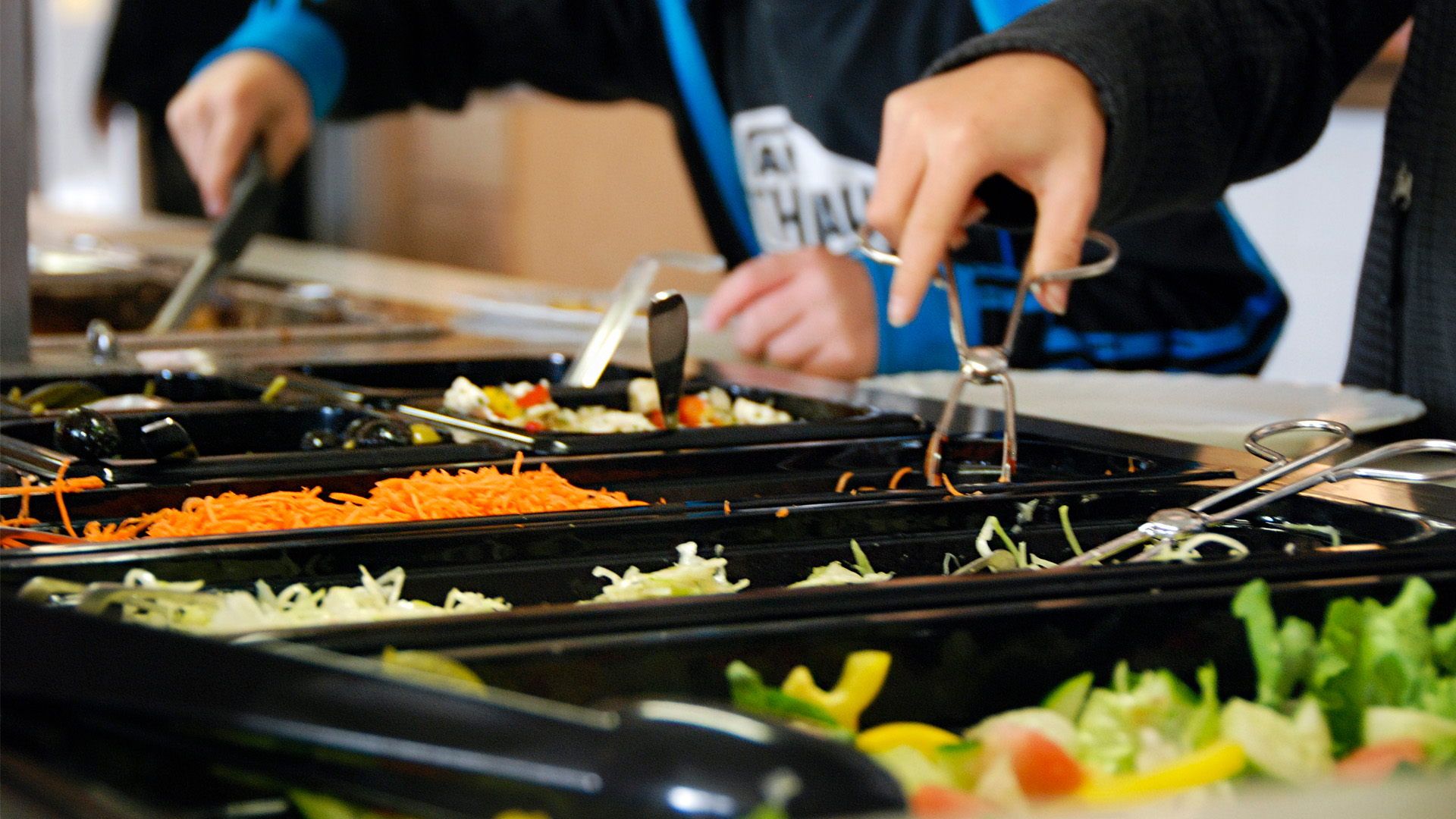 Två personer tar sallad i en salladsbuffé.