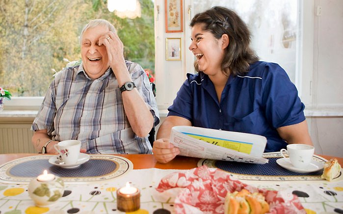 Äldre herre får besök av kvinna från hemtjänsten, de sitter vid ett bort och skrattar.
