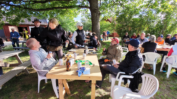 Människor som sitter vid stolar och bord under ett träd. Soligt väder.